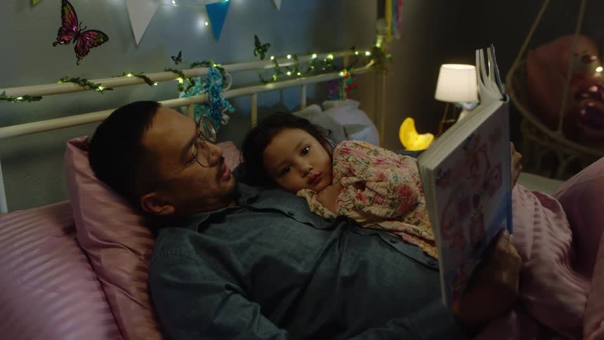 A father lying on the bed with his daughter on his chest reading her a bedtime story in the bedroom