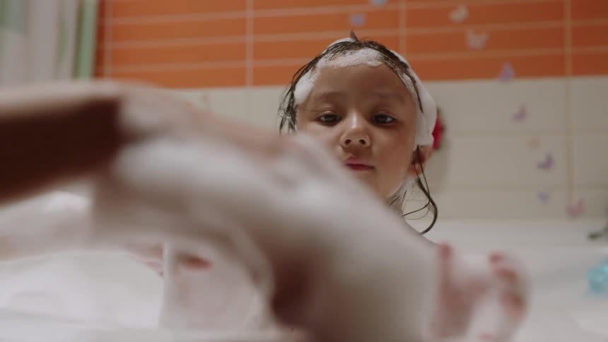 The video shows an adult and a young girl in a brightly lit bathroom taking a bubble bath the adult is playing with the girl creating large playful shapes with the foam the girl is smiling and appears to be enjoying the bath