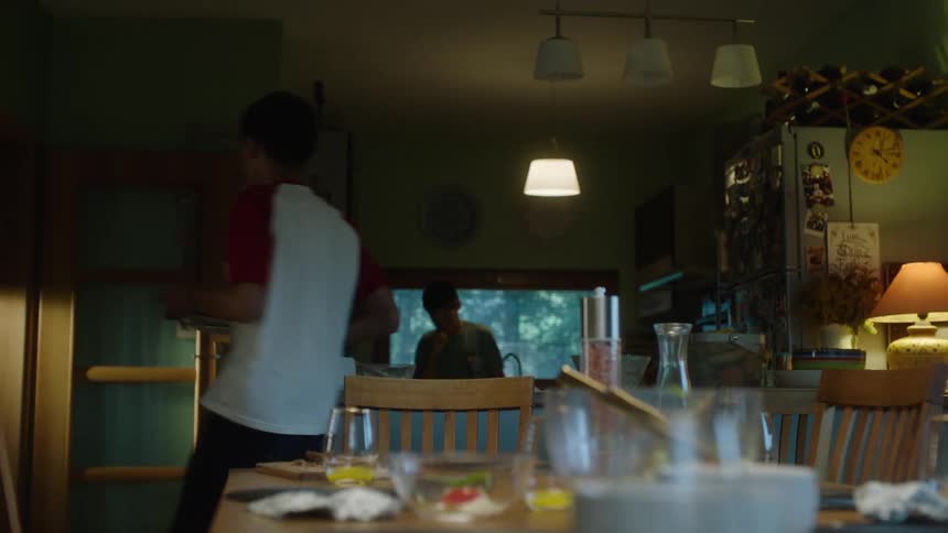 The video shows two people a man and a boy clearing a dinner table in a kitchen or dining area the man takes plates from the table while the boy picks up a bowl of salad with tongs and places it on a shelf