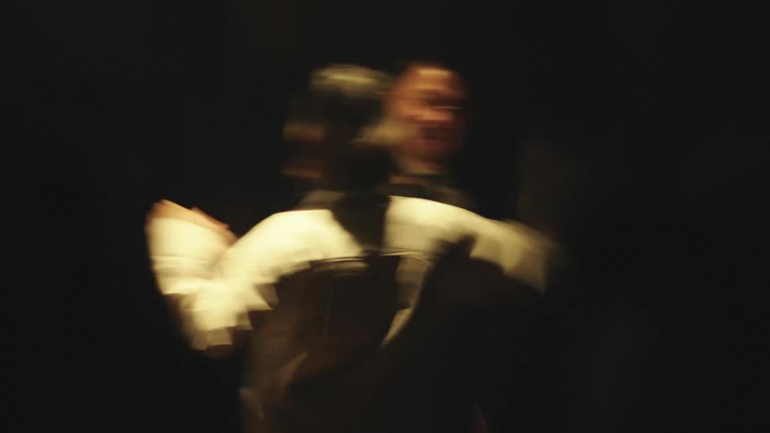 In a dimly lit room a woman in a white shirt and a man with suit and glasses are dancing and having an intimate moment in a time lapse capture