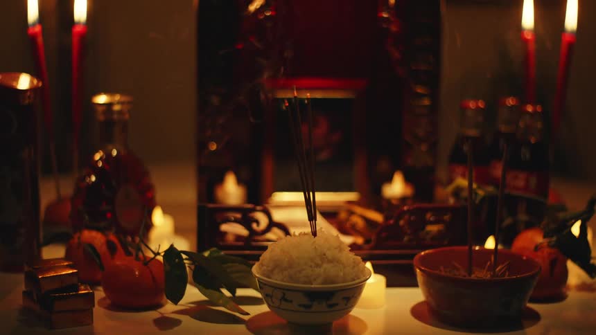 A traditional asian ritual temple with redlit candles surrounding a picture of a deceased woman as someone places incense sticks into a bowl of rice