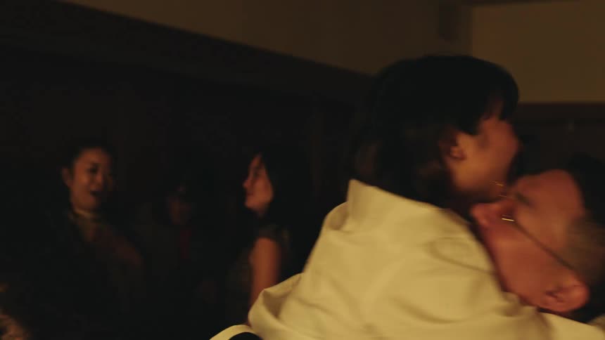 A young man wearing glasses walks behind a woman in a crowded room and she immediately hugs him excitedly upon seeing him