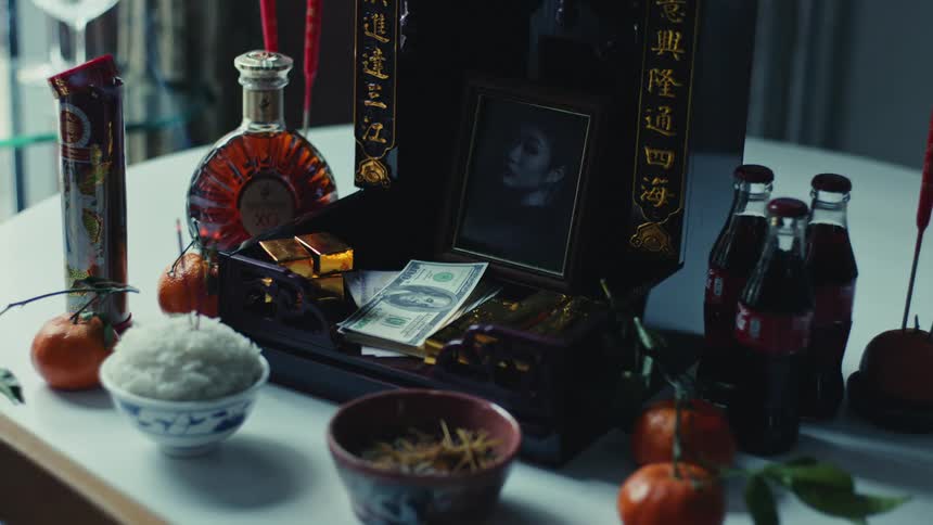 An altarlike setup with offerings including a bottle of liquor oranges rice and money along with a framed picture and other decorative items with chinese writing in the background