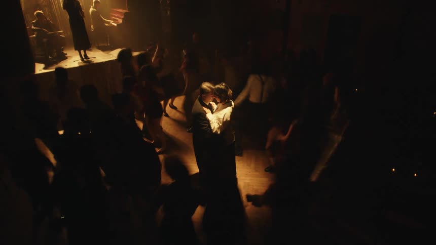 A couple hugging on a club stage while others around them dance to the music played by the singers