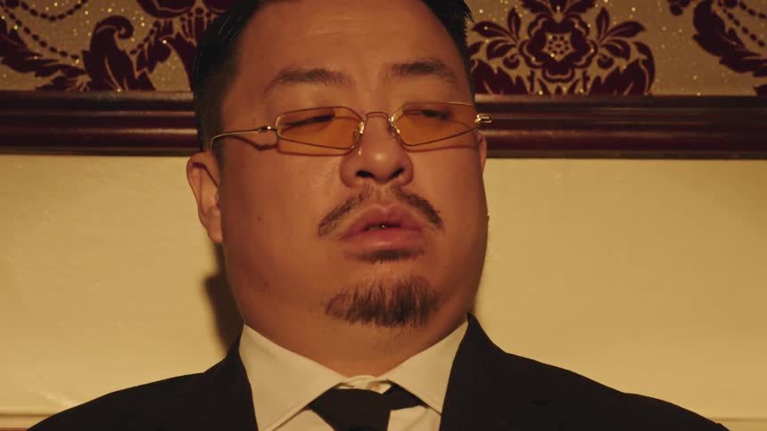 Man in a suit with gold glasses sits tiredly in a dimly lit room