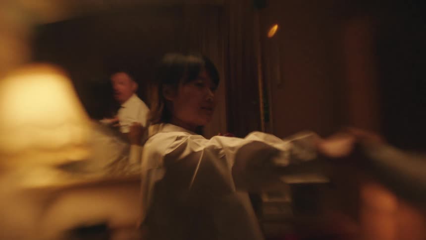 A man and a woman are dancing closely in a dimly lit room with the woman playfully throwing her arms up while the man smiles and looks at her