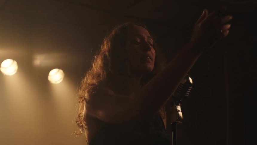 A woman stands under warm stage lights gesturing with her hands while singing into a microphone