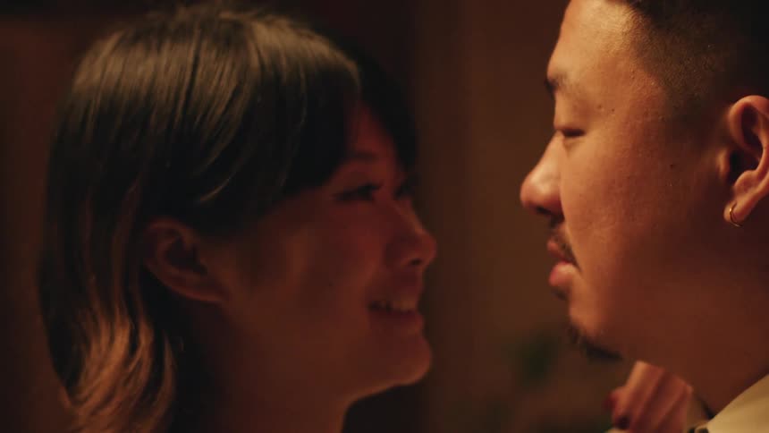 In a dark room a man and a woman stand in close proximity to each other sharing an intimate romantic moment as they smile and laugh