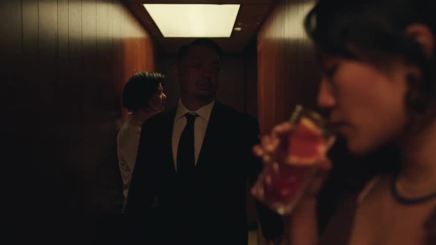 A man wearing a black suit walking throughout a hall and there are two women drinking something