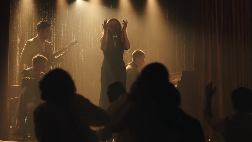 A female singer is performing on a stage with a band bathed in spotlights and facing an audience in a darkened music venue