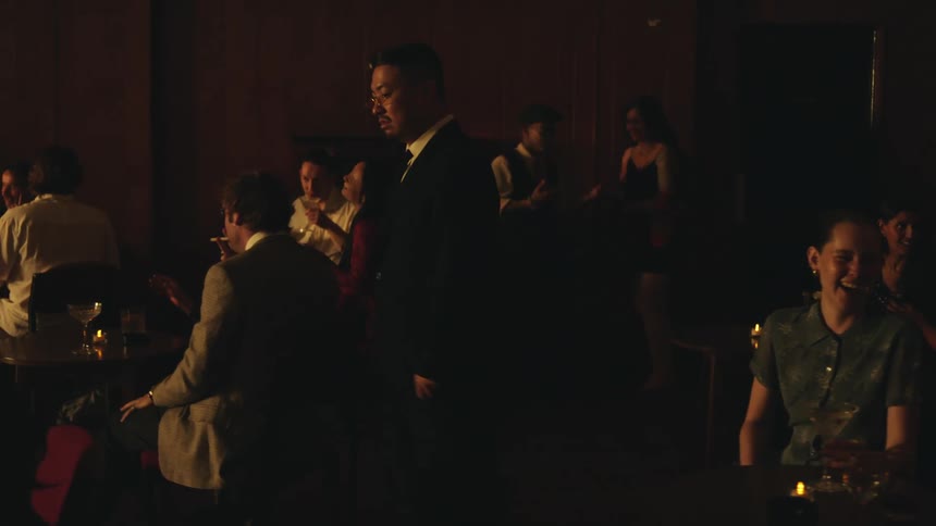 A man wearing a suit and glasses walks into a dimly lit room which is filled with other people socializing and dancing and he looks around at the scene