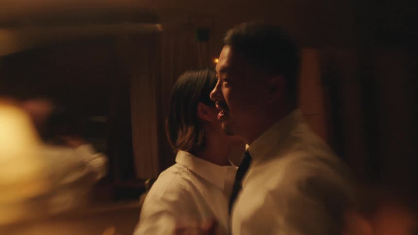 A man and a woman in white shirts dance in a dimly lit warm toned room their reflections visible in a mirror behind them