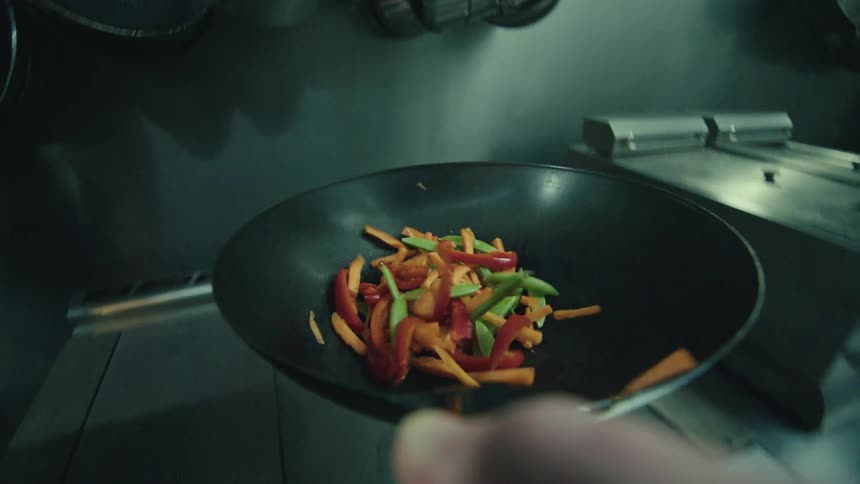 A person is shown expertly stirfrying julienned red peppers and snap peas in a large wok tossing them into the air in a kitchen environment