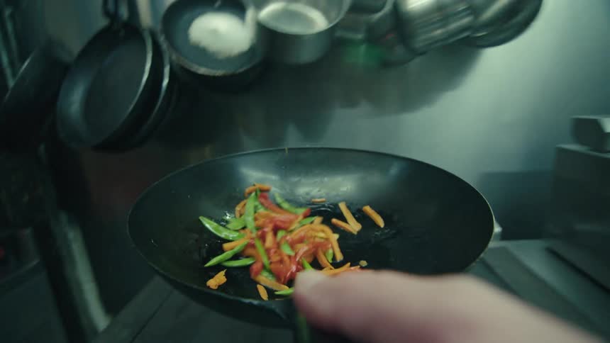 A closeup shot shows a professional chefs hand using a ladle to stirfry red and green vegetables in a black wok with the motion of the food being tossed and flipped clearly visible in the foreground