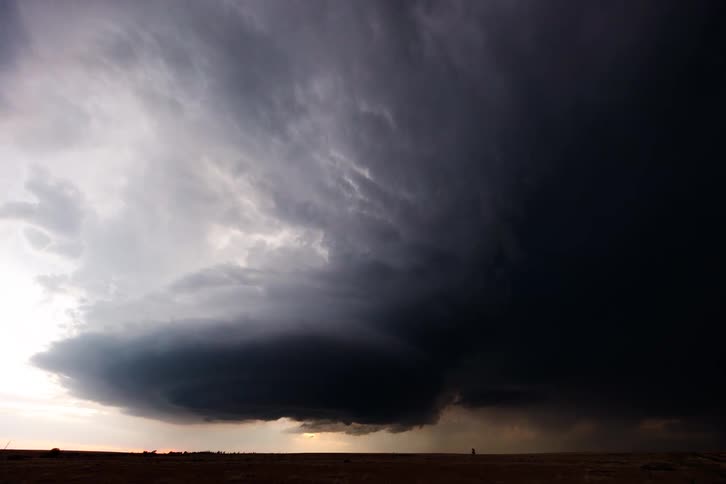 A large dark storm cloud with a textured and swirling surface hangs over a flat brown landscape with bright flash of lightning on the right