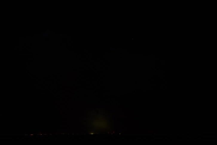 The video shows a dark night sky with flashes of lightning inside a cloud the horizon is visible in the lower portion of the frame where a small faintly lit cityscape is barely visible