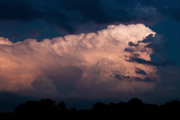 A huge cumulonimbus cloud is illuminated by the late afternoon sun glowing with shades of pink and orange the dark blue sky surrounds the massive cloud while the silhouettes of trees can be seen at the bottom of the frame
