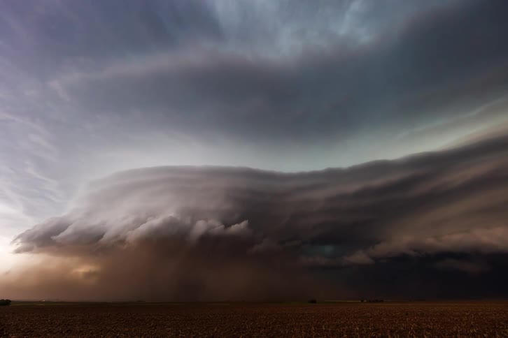 A massive dark and rapidly rotating supercell thunderstorm moving ominously across a flat open field with a wall of dust and rain being kicked up at the base of the storm