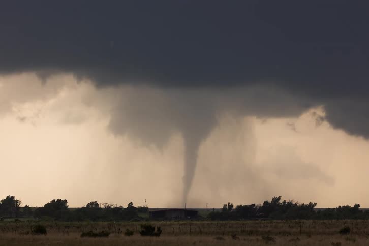A tornado touches down in the middle of a large field with a funnel cloud descending from a dark gray storm cloud a small white house sits below the swirling vortex while the sky in the background has an ominous yellow glow