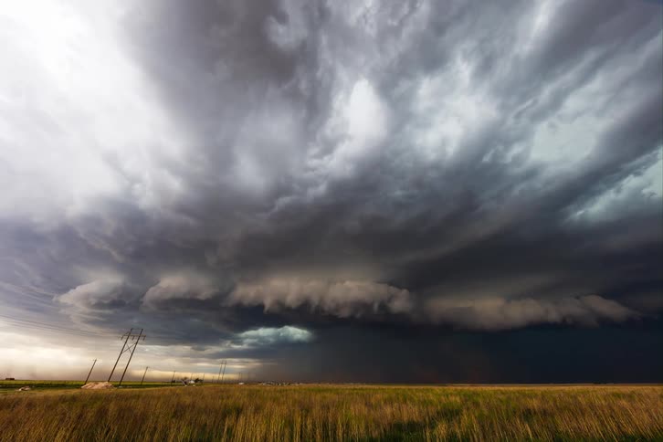 A vast ominous storm cloud blankets the sky with its dark chaotic shape dominating the frame the cloud extends downward almost touching the ground where the rain and wind can be seen kicking up dust from the dry field