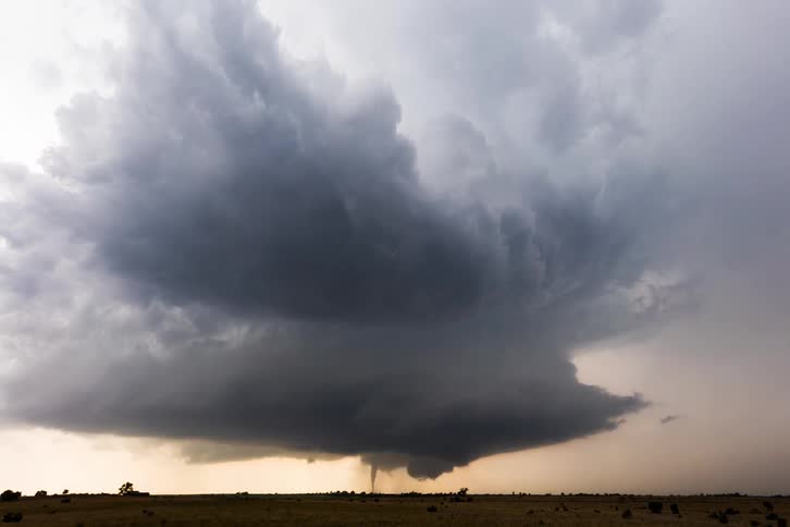 A dark and ominous supercell thunderstorm with a tornado forming at its base moving across a flat open field