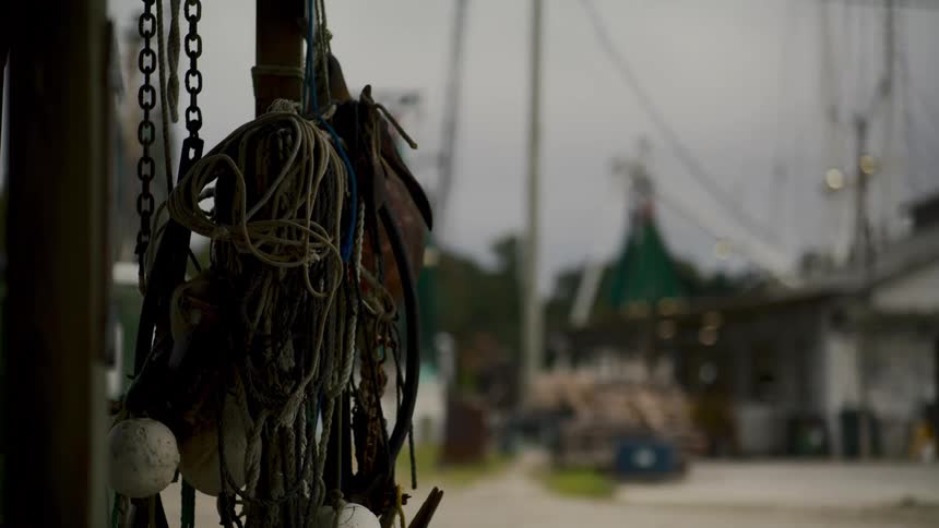 A collection of weathered ropes chains and buoys hang from a pole in the foreground of a blurry outoffocus shot of a boat dock with old fishing vessels visible in the background