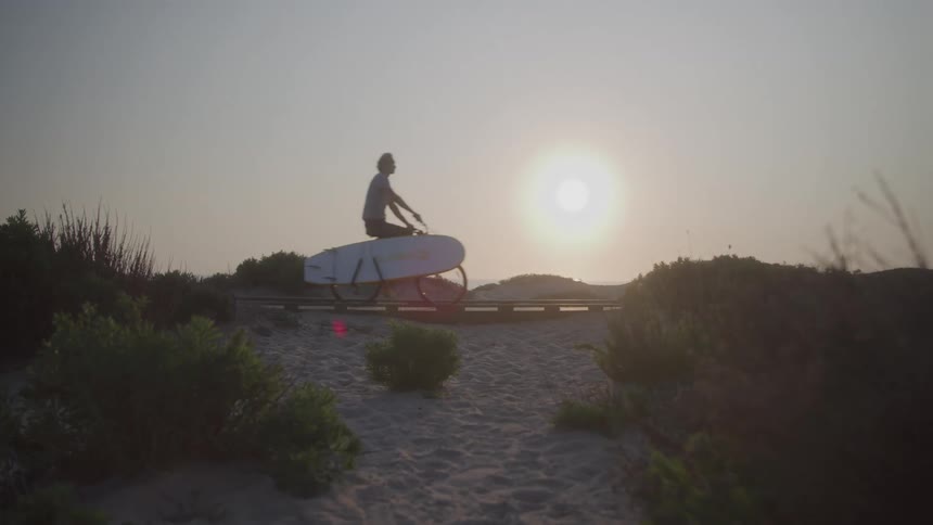 A person riding a bicycle with a surfboard on a sandy path with the sun visible in the background