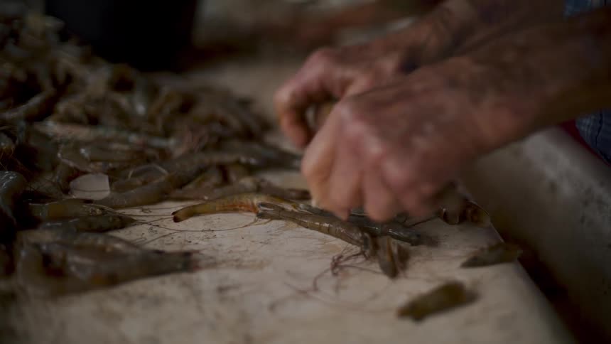 Two people stand side by side using their hands to quickly select and clean shrimp on a light brown surface