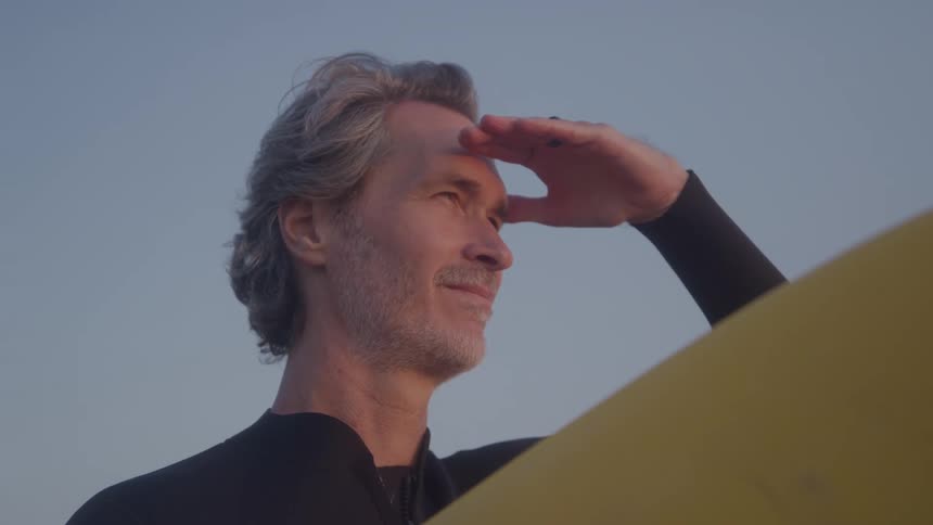 An older man in a wetsuit holding a yellow surfboard as he shields his eyes with his hand while looking out