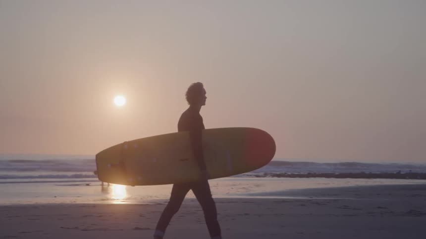 A surfer silhouetted against a setting sun walks along a beach carrying a yellow surfboard under their arm