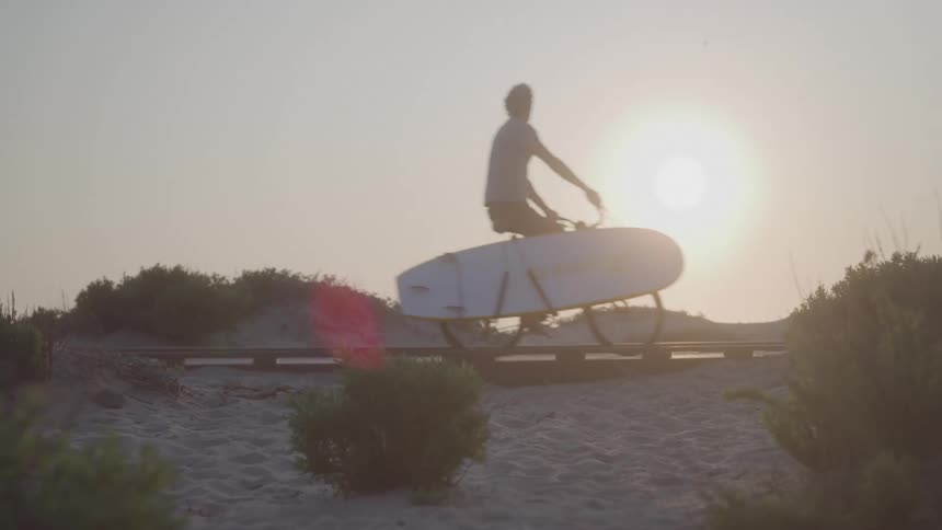 A man on a bicycle with a surfboard attached to the bicycle goes over a wood path on a beachs sand