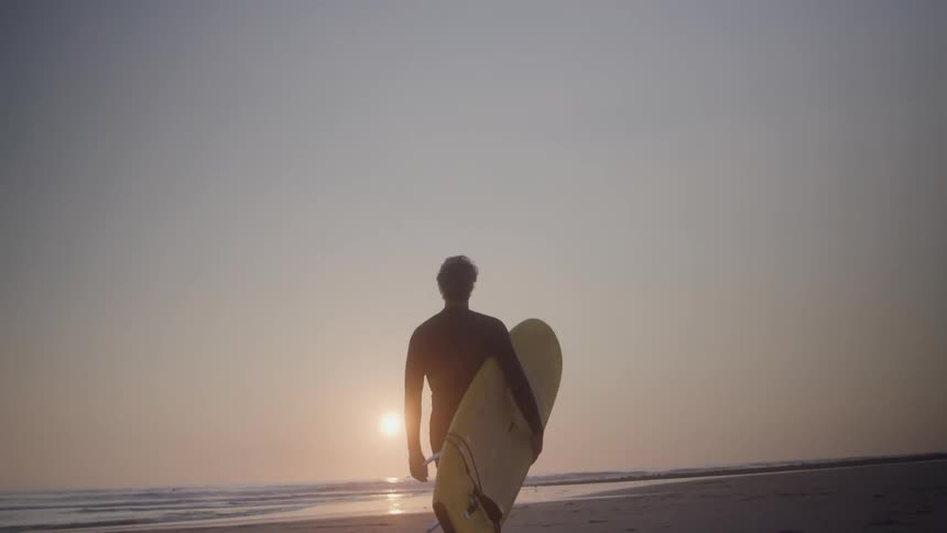 A mans silhouette holding a yellow surfboard standing on a beach with his back to the camera with the bright sun low on the horizon