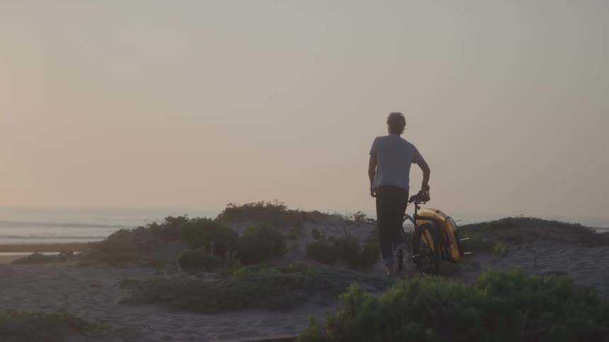 Man carrying a bicycle with a surfboard on it and walking towards the beach outdoors during dusk or dawn