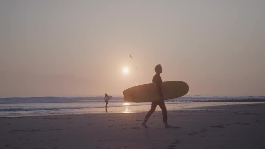 A surfer walks on a beach with his board under his arm parallel to the sea as another surfer also carrying his board heads towards the water
