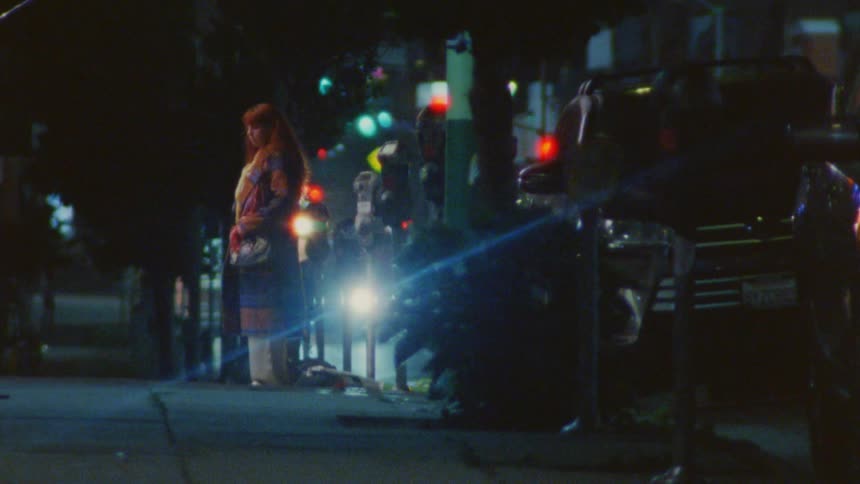 A woman standing on the sidewalk by the road at night puts down her phone from her ear with a frustrated expression