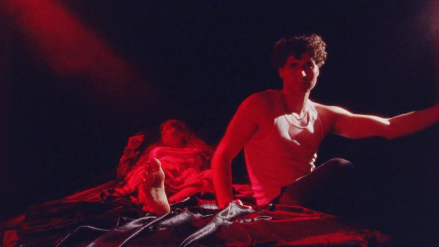 A woman lying in bed and a man sitting on the edge of the bed with a raised arm neither of them moving with the room flooded with red light
