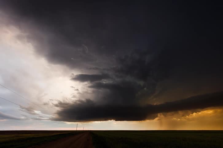 A powerful and massive supercell storm cloud forming and darkening in the sky above a flat empty landscape with a dirt road