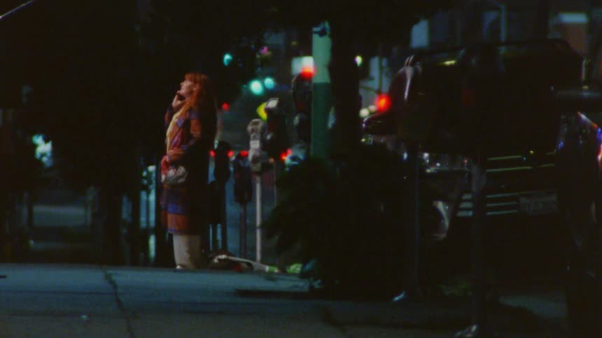 Redhead woman standing on the sidewalk next to parked cars on a suburban area talking on a phone outdoors during night time