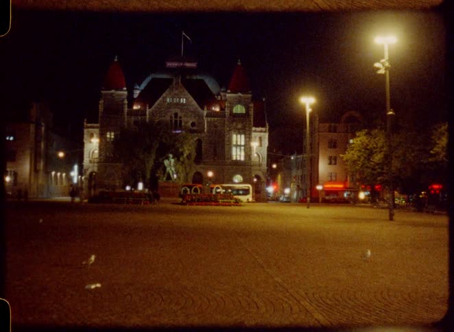 City bus stopping at a bus station between an old building and a park with pedestrian walking around outdoors during night time