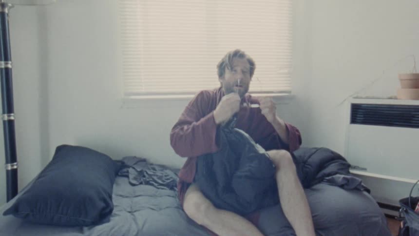 White man wearing glasses and a robe waking up in a bed and searching for his glasses indoors during day time