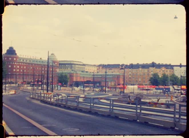 A wide view of a city square with historical brick buildings where traffic is moving around a central roundabout and trams run on their tracks