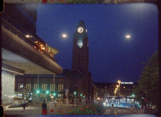 A busy street scene at night shows a prominent clock tower in the distance and vehicles driving by all under a blue sky illuminated by bright lights