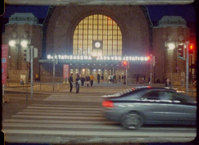 The video shot with a retro grainy film effect shows a street scene at night in front of a grand building with a large arched window the camera is static capturing cars driving by and people walking across the crosswalk