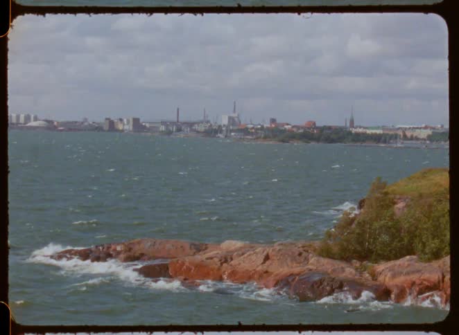 A rocky shoreline being hit by sea waves with a city visible across the ocean at midday
