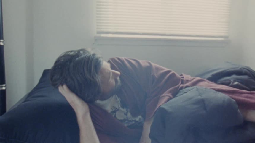 A middleaged man being woken up from sleep by someone else putting on his glasses at midday