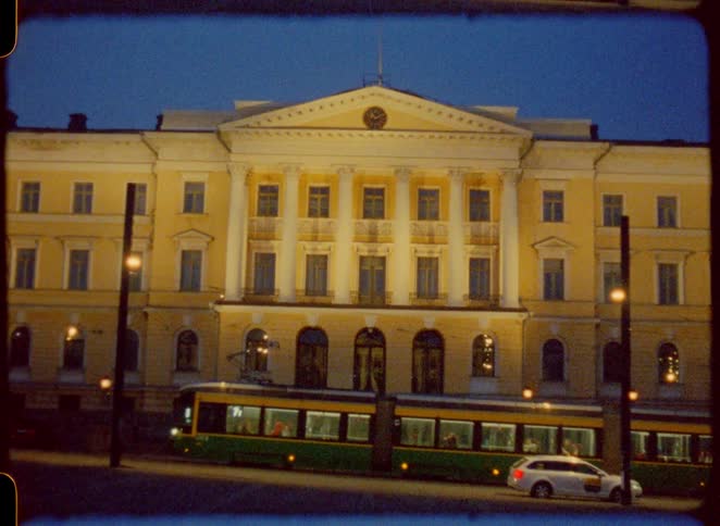 A building with many windows and an arched entryway is shown at night with a tram driving past in a city setting