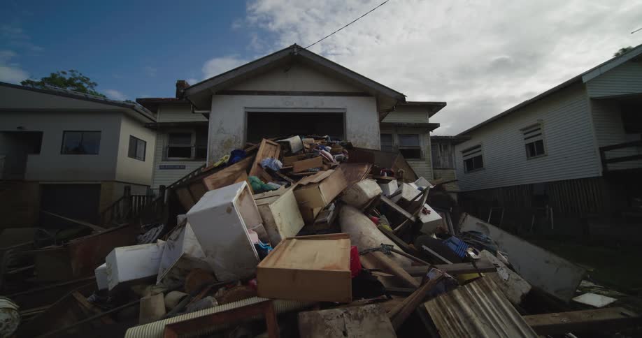 A wide shot of a house shows a large pile of debris and furniture stacked up in the front yard a man in a highvisibility vest walks over to the pile and begins to sort through it