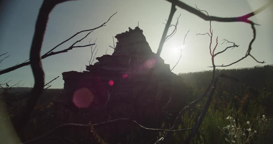 Silhouette of a rugged crumbling stone monument against a bright hazy sky with sunlight flaring around its edges and dried branches framing the shot