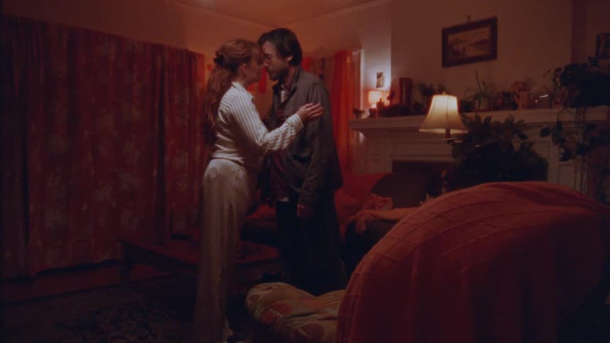 A couple standing close together in a dimly lit cozy living room sharing a tender and intimate moment as they embrace and move in for a kiss