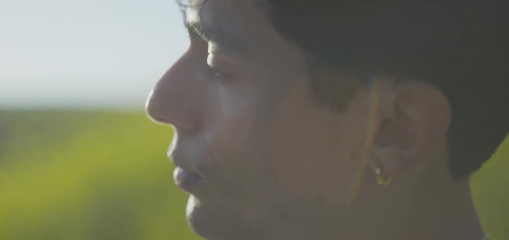 A mans face with a neutral and pensive expression as he looks to the side in a sunny outdoor setting with a blurred yellowgreen background
